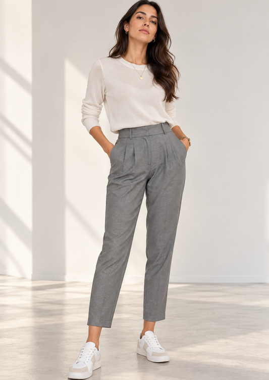 A woman modeling a pair of high-waisted, light gray pleated trousers with a white long-sleeve top and white sneakers, standing in a bright, minimalist room with soft window shadows.